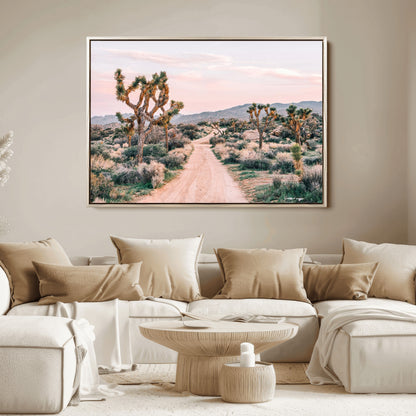 A dusty road winding through Joshua Tree National Park, framed by iconic twisted trees and warm desert tones under a soft pastel sky.