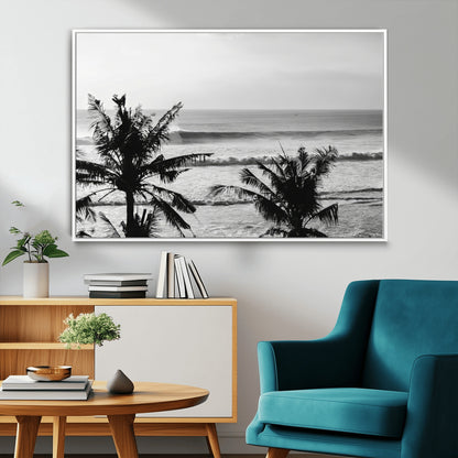 Black and white view of ocean waves crashing on a tropical coastline, framed by silhouetted palm trees under a vast, open sky.