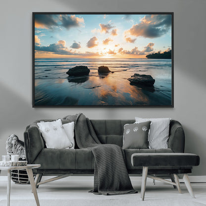 A serene beach at sunset with vibrant clouds, calm water, and rocks in the foreground reflecting the warm colors of the setting sun.