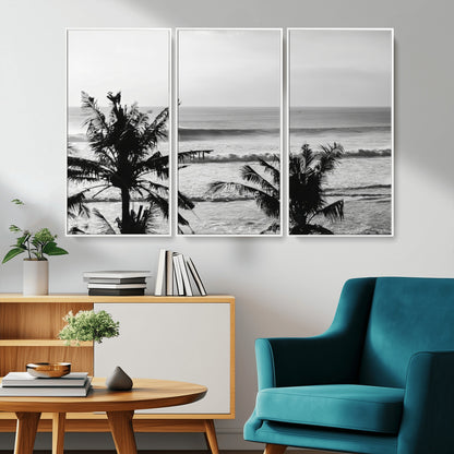 Black and white view of ocean waves crashing on a tropical coastline, framed by silhouetted palm trees under a vast, open sky.