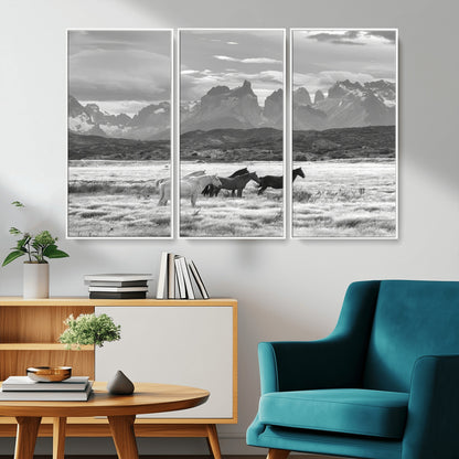 A black and white landscape of wild horses grazing in a vast grassland with towering mountains in the background, creating a moody and rustic western aesthetic.