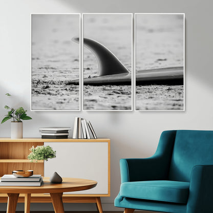 A black and white close-up of a surfboard fin resting on the sandy shore, highlighting minimalist beach aesthetics and classic surf culture.