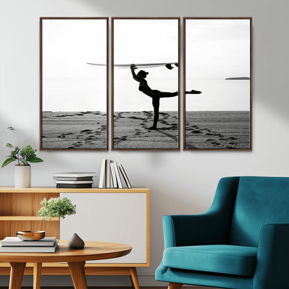 A woman balances a surfboard on her head while striking a yoga pose on the beach, silhouetted against a calm ocean horizon.