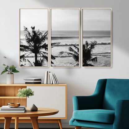 Black and white view of ocean waves crashing on a tropical coastline, framed by silhouetted palm trees under a vast, open sky.