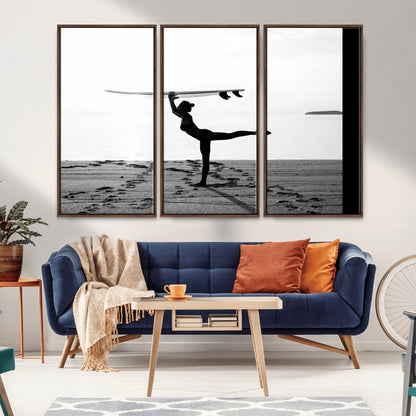 A woman balances a surfboard on her head while striking a yoga pose on the beach, silhouetted against a calm ocean horizon.
