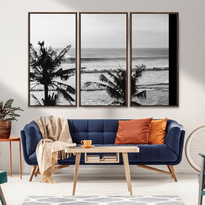 Black and white view of ocean waves crashing on a tropical coastline, framed by silhouetted palm trees under a vast, open sky.