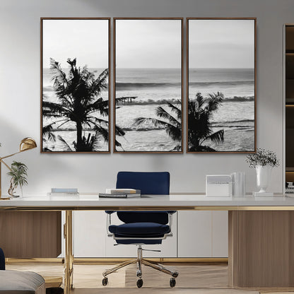 Black and white view of ocean waves crashing on a tropical coastline, framed by silhouetted palm trees under a vast, open sky.