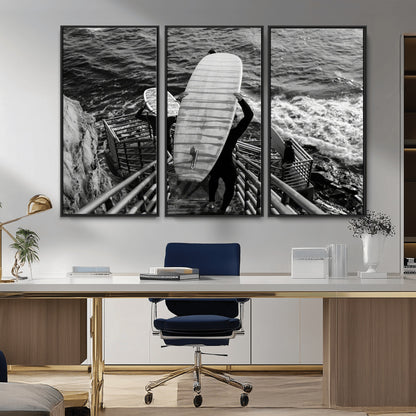 Black and white photo of surfers carrying boards down a rugged cliffside stairway, capturing a bold and adventurous coastal scene.