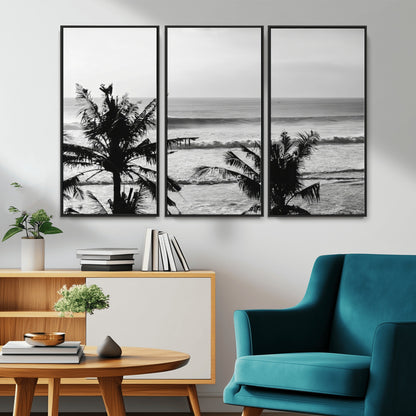 Black and white view of ocean waves crashing on a tropical coastline, framed by silhouetted palm trees under a vast, open sky.