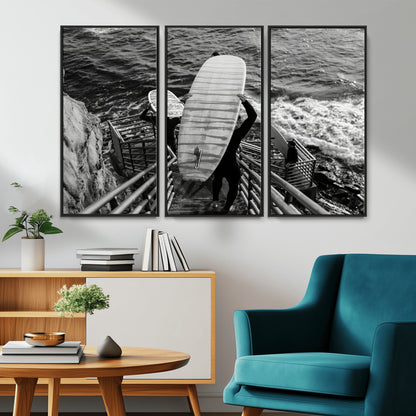 Black and white photo of surfers carrying boards down a rugged cliffside stairway, capturing a bold and adventurous coastal scene.