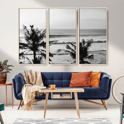 Black and white view of ocean waves crashing on a tropical coastline, framed by silhouetted palm trees under a vast, open sky.