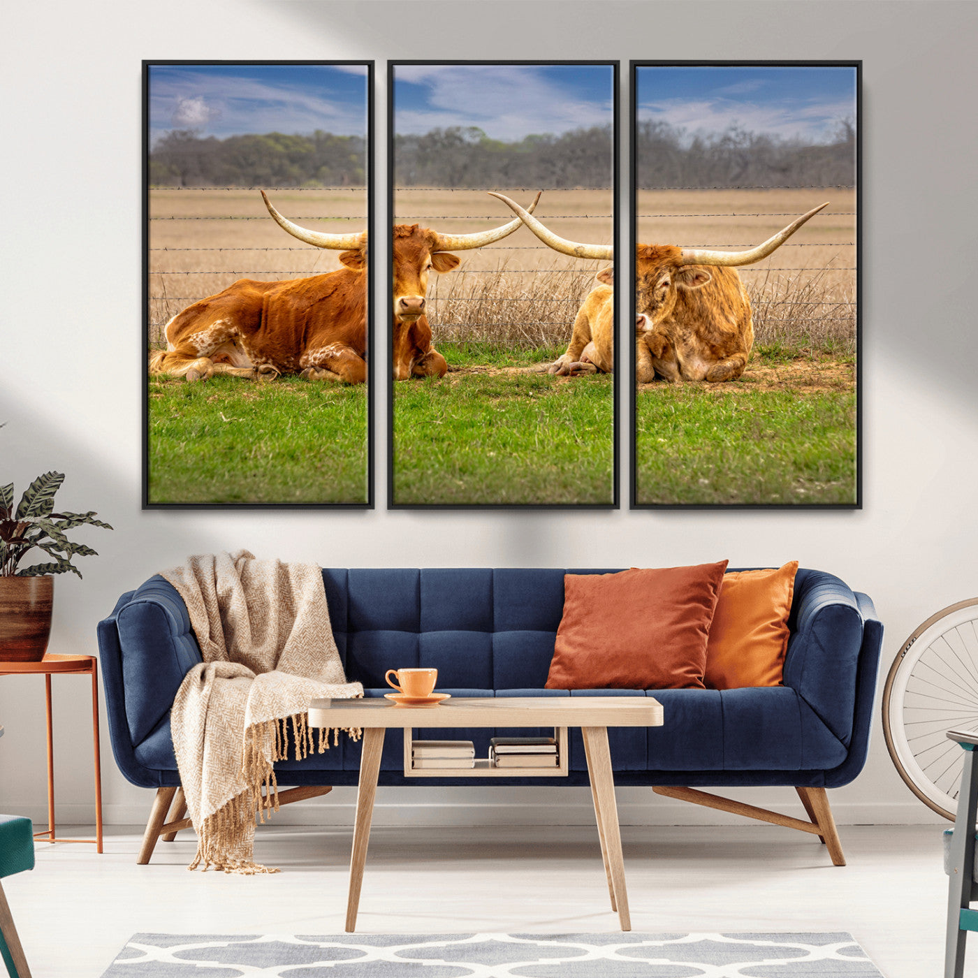 Two longhorn cows lying on green grass in front of a golden field and trees under a blue sky, perfect for rustic country wall decor.