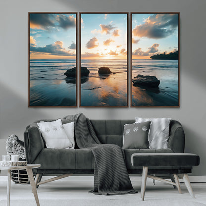 A serene beach at sunset with vibrant clouds, calm water, and rocks in the foreground reflecting the warm colors of the setting sun.