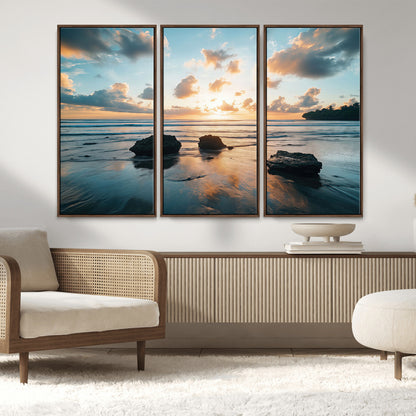 A serene beach at sunset with vibrant clouds, calm water, and rocks in the foreground reflecting the warm colors of the setting sun.