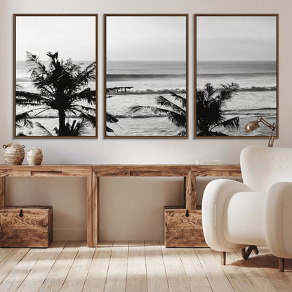 Black and white view of ocean waves crashing on a tropical coastline, framed by silhouetted palm trees under a vast, open sky.