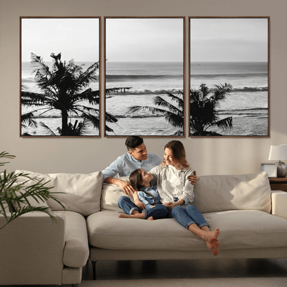Black and white view of ocean waves crashing on a tropical coastline, framed by silhouetted palm trees under a vast, open sky.