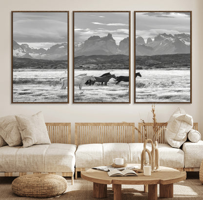 A black and white landscape of wild horses grazing in a vast grassland with towering mountains in the background, creating a moody and rustic western aesthetic.