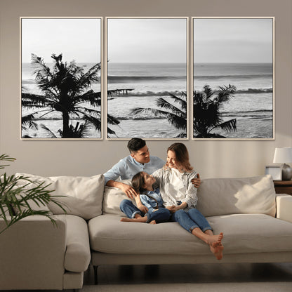 Black and white view of ocean waves crashing on a tropical coastline, framed by silhouetted palm trees under a vast, open sky.
