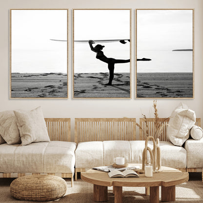 A woman balances a surfboard on her head while striking a yoga pose on the beach, silhouetted against a calm ocean horizon.