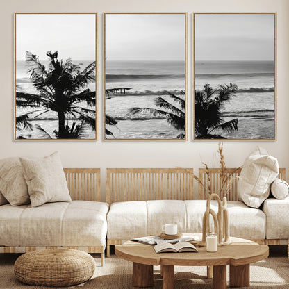 Black and white view of ocean waves crashing on a tropical coastline, framed by silhouetted palm trees under a vast, open sky.