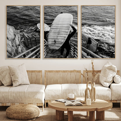 Black and white photo of surfers carrying boards down a rugged cliffside stairway, capturing a bold and adventurous coastal scene.