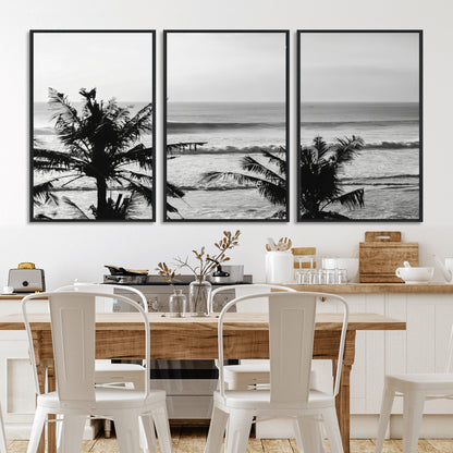 Black and white view of ocean waves crashing on a tropical coastline, framed by silhouetted palm trees under a vast, open sky.