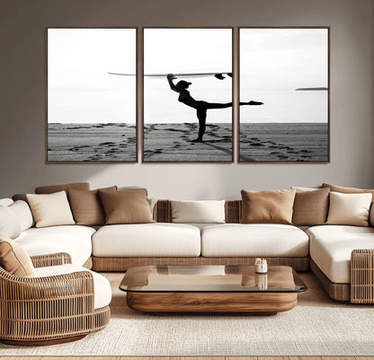 A woman balances a surfboard on her head while striking a yoga pose on the beach, silhouetted against a calm ocean horizon.