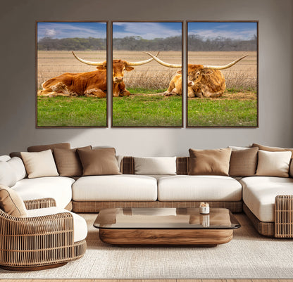 Two longhorn cows lying on green grass in front of a golden field and trees under a blue sky, perfect for rustic country wall decor.