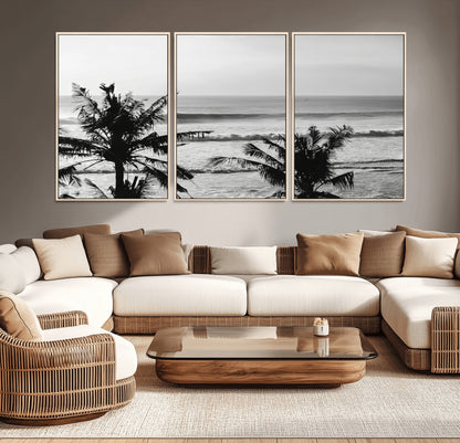 Black and white view of ocean waves crashing on a tropical coastline, framed by silhouetted palm trees under a vast, open sky.