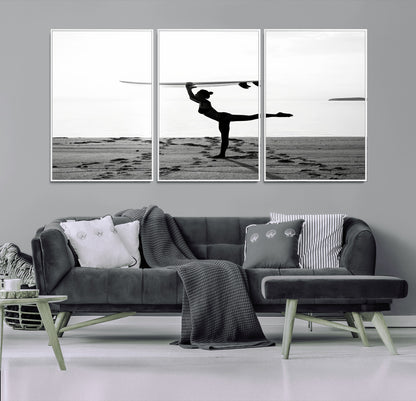 A woman balances a surfboard on her head while striking a yoga pose on the beach, silhouetted against a calm ocean horizon.