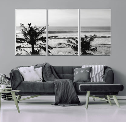 Black and white view of ocean waves crashing on a tropical coastline, framed by silhouetted palm trees under a vast, open sky.