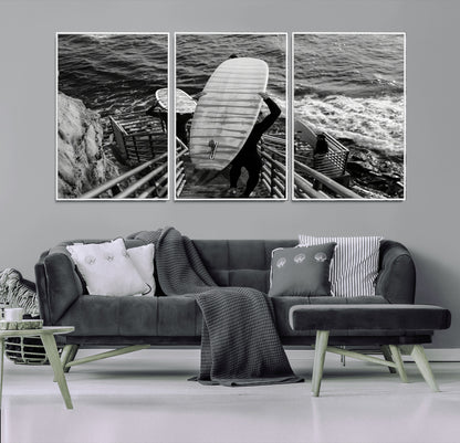 Black and white photo of surfers carrying boards down a rugged cliffside stairway, capturing a bold and adventurous coastal scene.