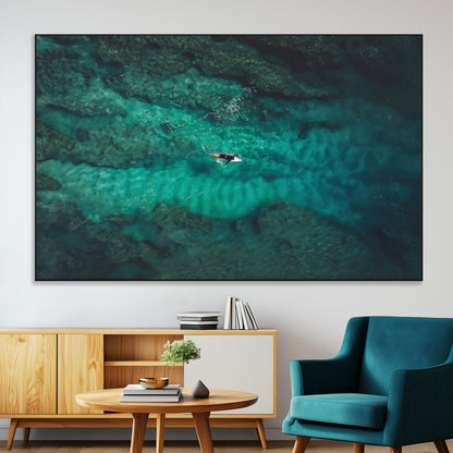 An aerial view of a solo surfer paddling through crystal-clear turquoise waters over a sunlit reef, capturing the beauty and serenity of open water surfing.
