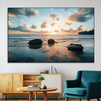 A serene beach at sunset with vibrant clouds, calm water, and rocks in the foreground reflecting the warm colors of the setting sun.