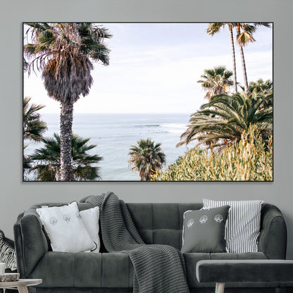 Scenic coastal view with tall palm trees overlooking a calm ocean, soft blue waves, and surfers in the distance, creating a peaceful tropical vibe.