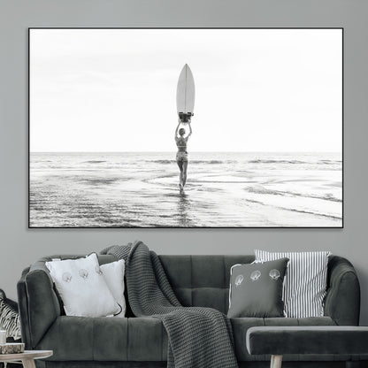 Black and white image of a female surfer holding a surfboard above her head while walking toward the ocean, creating a minimalist and bold coastal aesthetic.