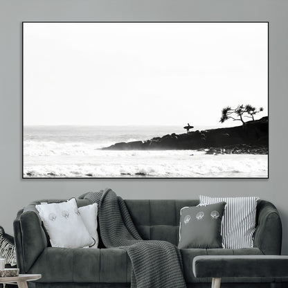 Black and white photo of a surfer standing on rocky cliffs, gazing at the waves, embodying the spirit of adventure and solitude.