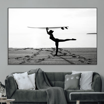 A woman balances a surfboard on her head while striking a yoga pose on the beach, silhouetted against a calm ocean horizon.