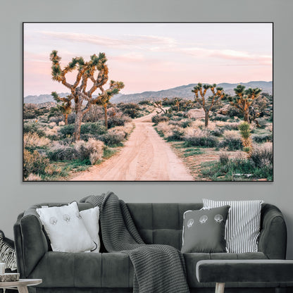 A dusty road winding through Joshua Tree National Park, framed by iconic twisted trees and warm desert tones under a soft pastel sky.