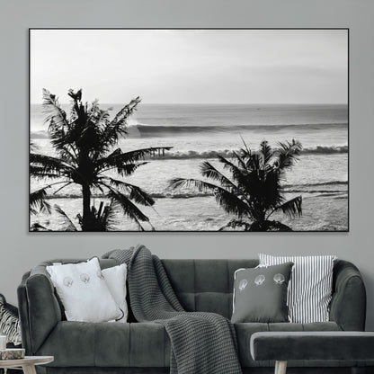 Black and white view of ocean waves crashing on a tropical coastline, framed by silhouetted palm trees under a vast, open sky.
