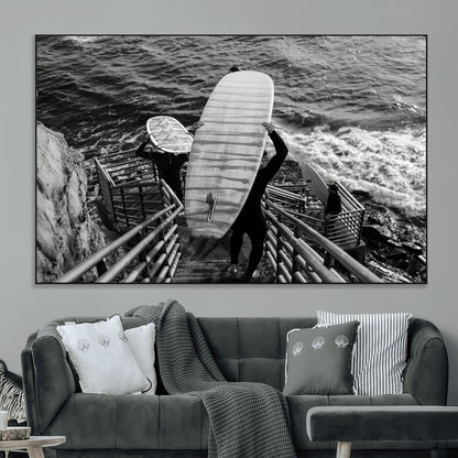 Black and white photo of surfers carrying boards down a rugged cliffside stairway, capturing a bold and adventurous coastal scene.