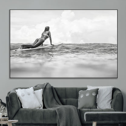 A black and white ocean scene featuring a surfer girl waiting on her board, gazing at the horizon, capturing a tranquil and adventurous coastal moment.