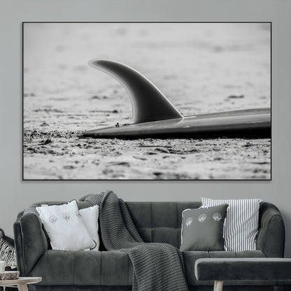 A black and white close-up of a surfboard fin resting on the sandy shore, highlighting minimalist beach aesthetics and classic surf culture.