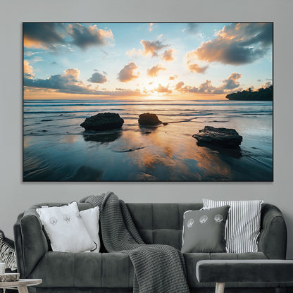 A serene beach at sunset with vibrant clouds, calm water, and rocks in the foreground reflecting the warm colors of the setting sun.