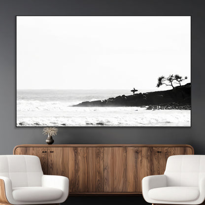 Black and white photo of a surfer standing on rocky cliffs, gazing at the waves, embodying the spirit of adventure and solitude.