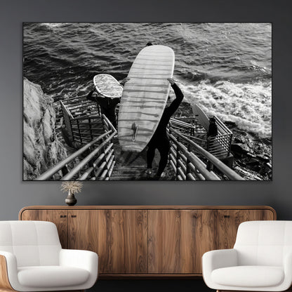 Black and white photo of surfers carrying boards down a rugged cliffside stairway, capturing a bold and adventurous coastal scene.
