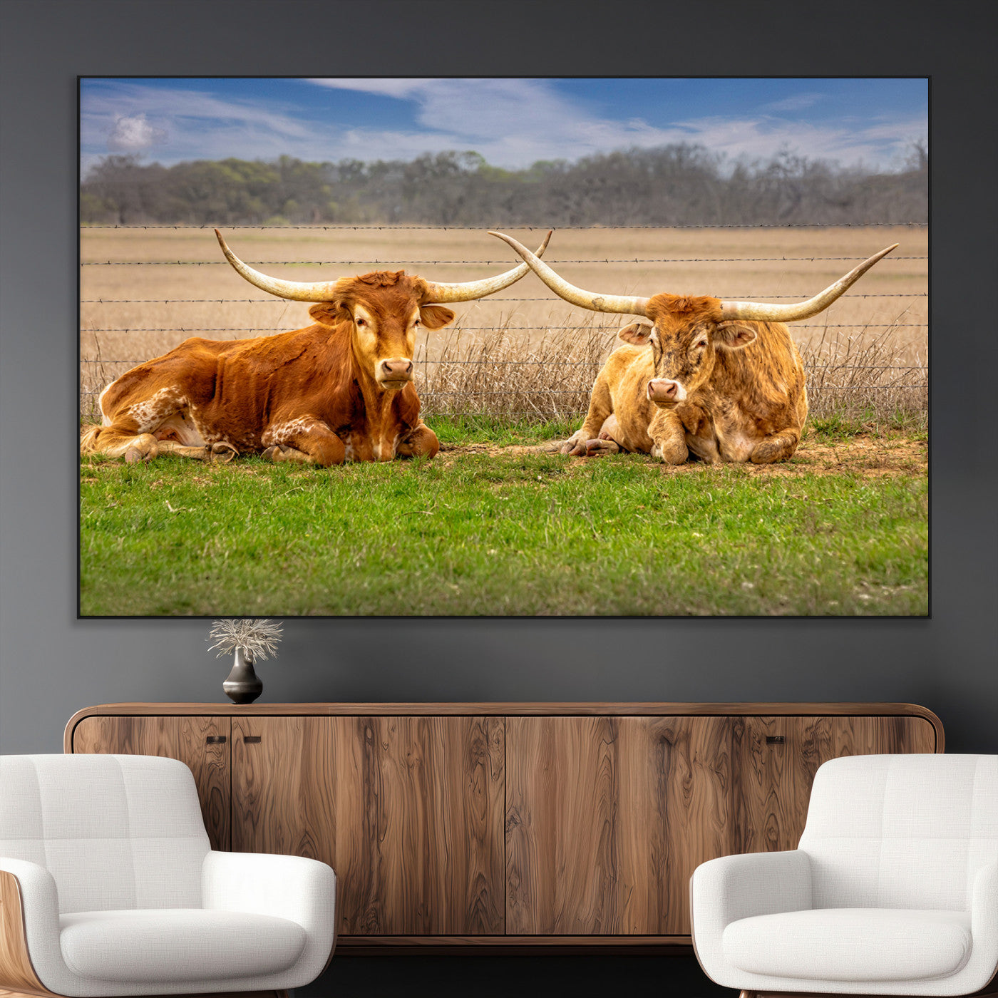 Two longhorn cows lying on green grass in front of a golden field and trees under a blue sky, perfect for rustic country wall decor.