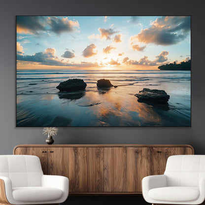 A serene beach at sunset with vibrant clouds, calm water, and rocks in the foreground reflecting the warm colors of the setting sun.