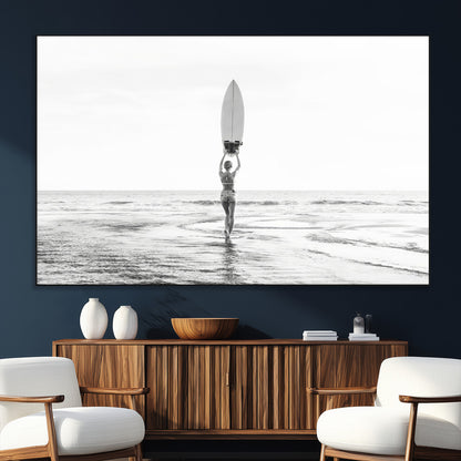 Black and white image of a female surfer holding a surfboard above her head while walking toward the ocean, creating a minimalist and bold coastal aesthetic.