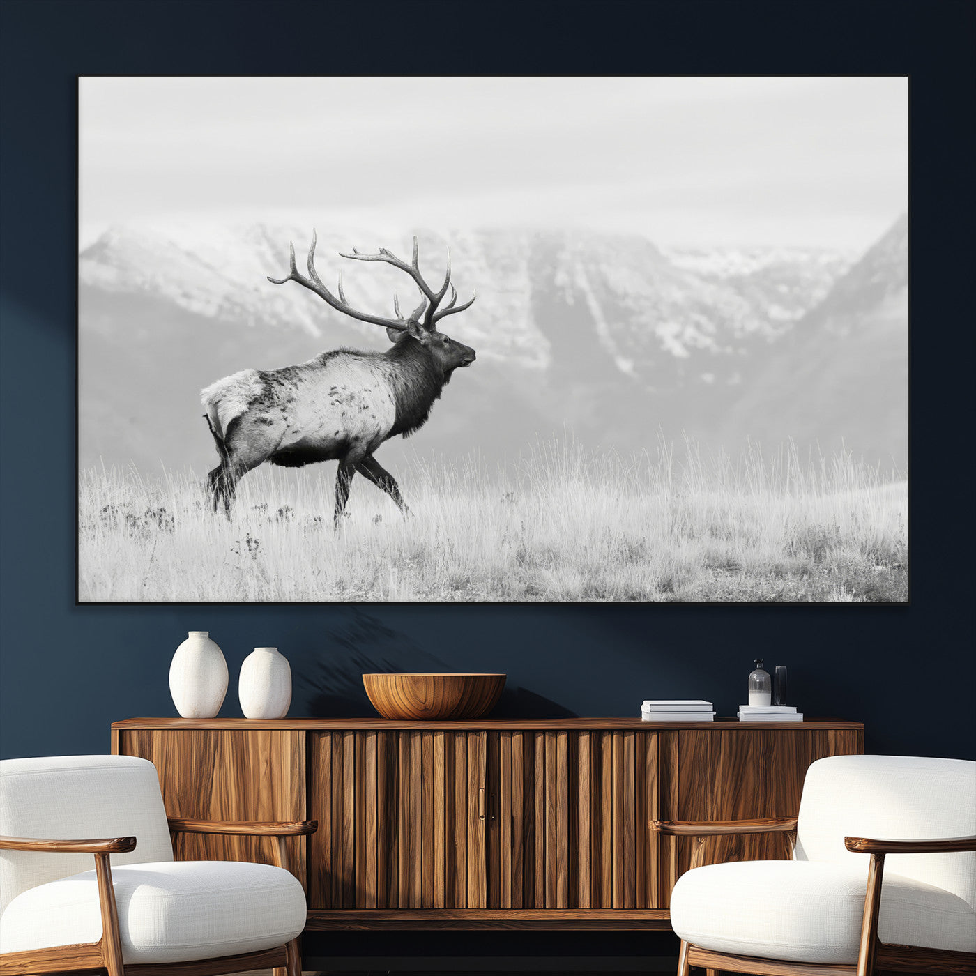 Black and white photograph of a majestic elk standing in a grassy field with snow-capped mountains in the background, creating a bold and rustic wilderness scene.